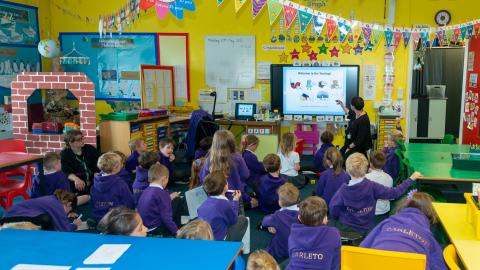 Year 2 pupils sat on the classroom floor , a teacher points at an interactive computer screen